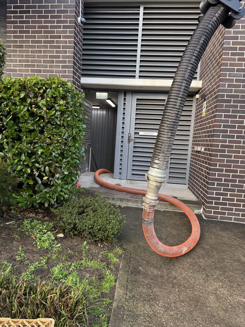 Suction hose routed past louvred plant-room doors beside a brick building