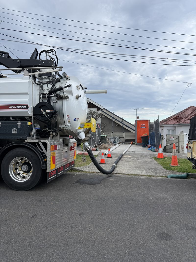 Vacuum truck on a residential street with its suction hose run up a driveway past traffic cones