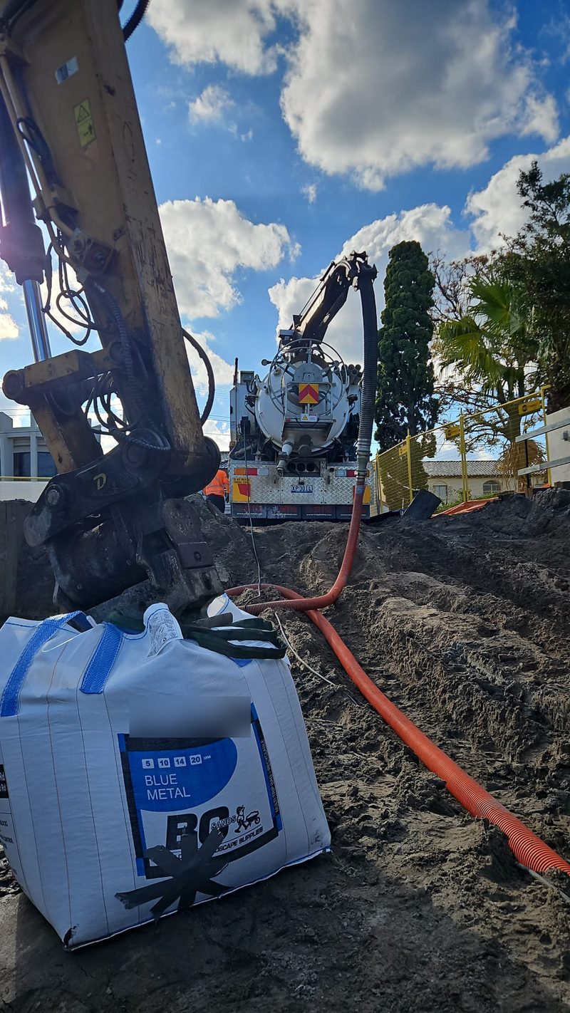 Excavator working alongside a vacuum truck with the suction hose run across an open excavation