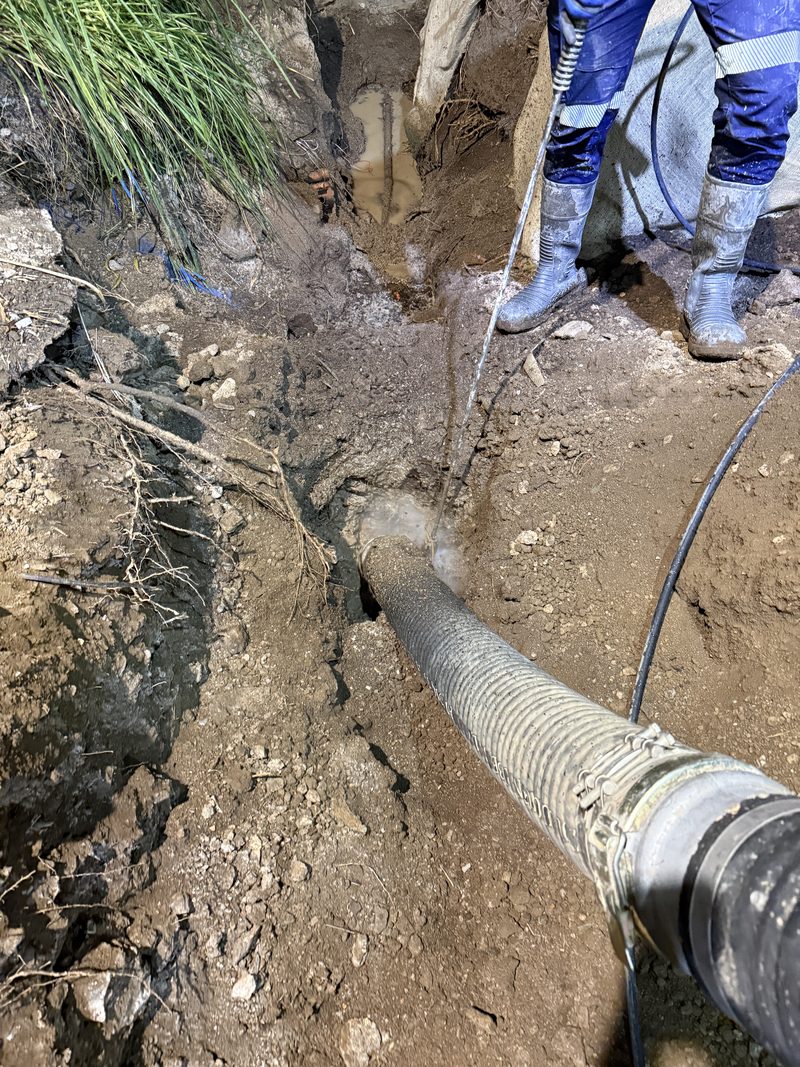 Worker using a water lance beside a suction hose in a trench with an exposed service line