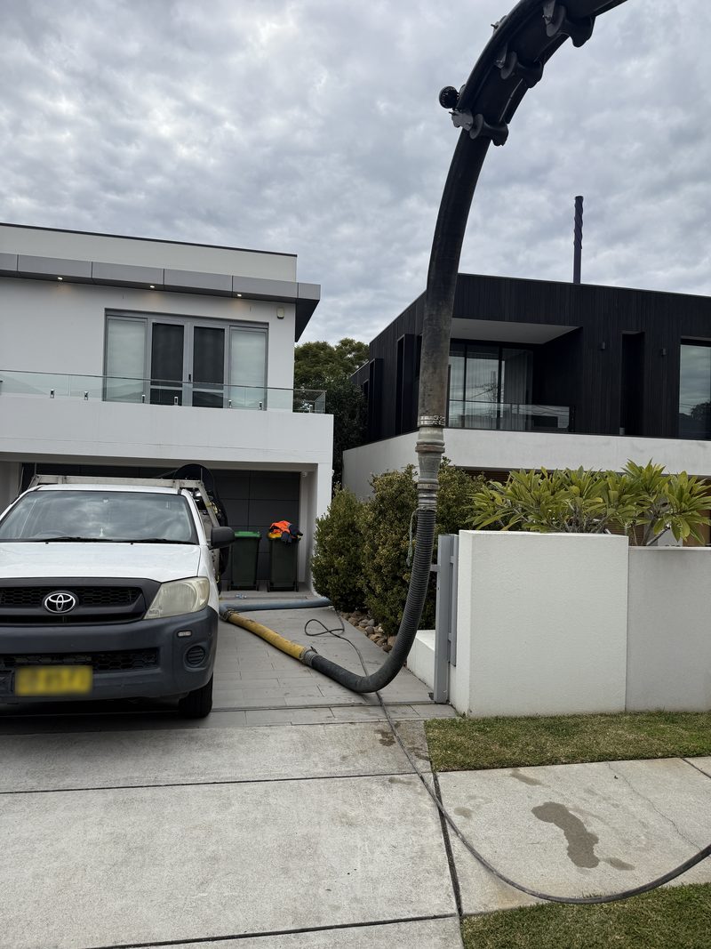 Vacuum truck boom reaching over a driveway with the suction hose run past a parked ute at a home
