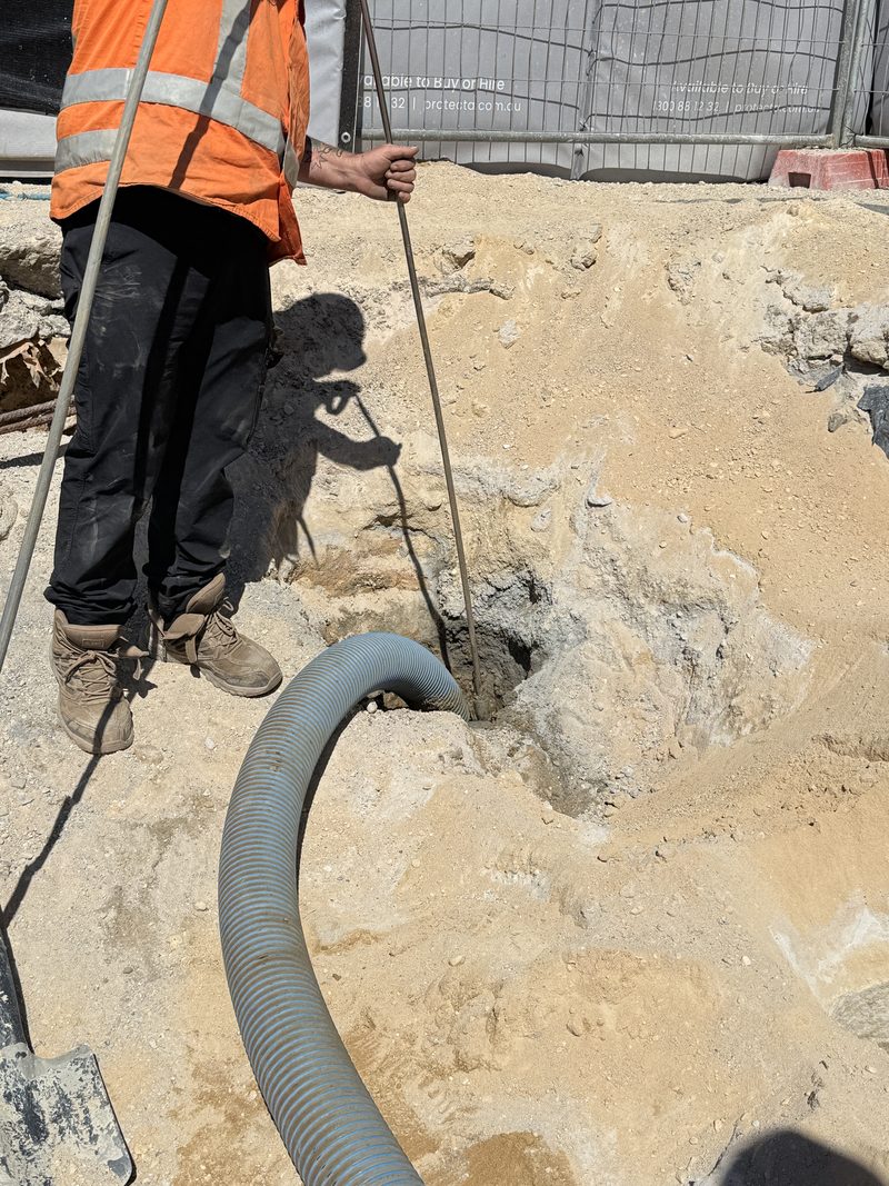 Worker probing a sandy excavation beside a suction hose on a fenced work site
