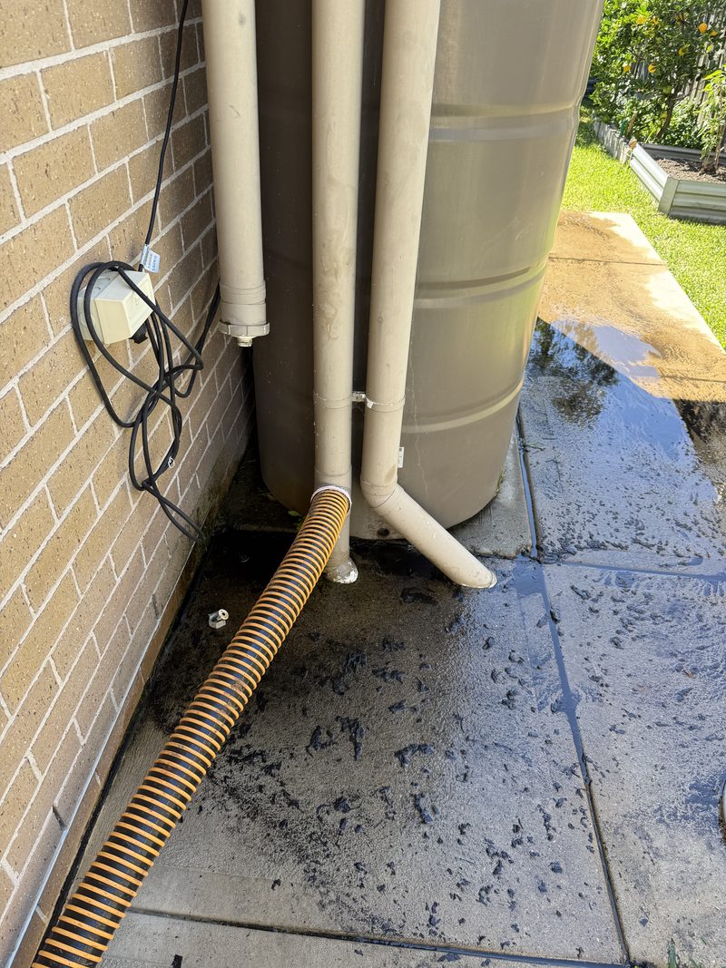 Suction hose run along a paved side path beside a rainwater tank at a home