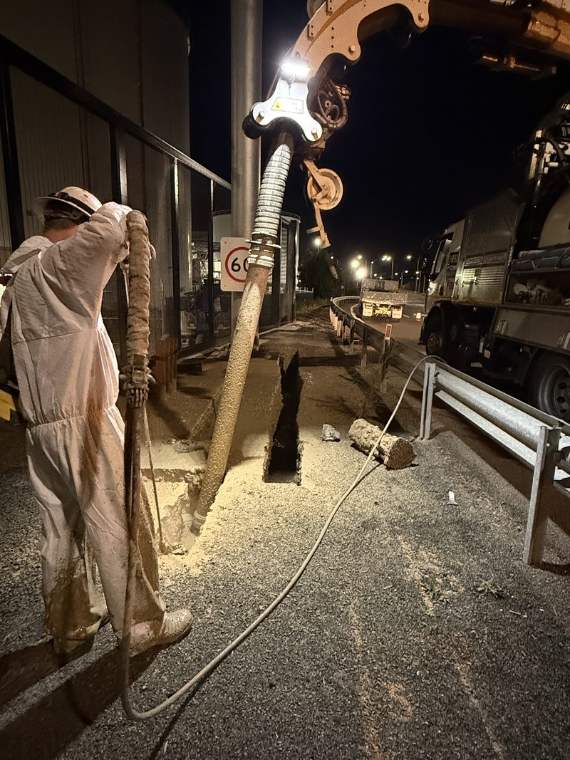 Worker guiding a suction hose and water lance into a narrow trench cut through asphalt beside a building at night
